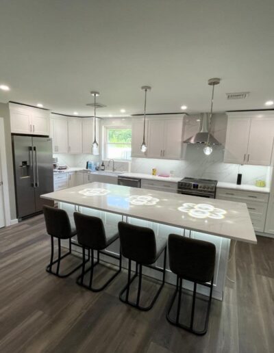 Modern kitchen with white cabinets, stainless steel appliances, and a large island with four black stools. The room has wood flooring and is lit by pendant lights and recessed lighting.