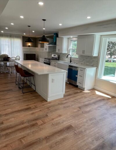 Modern kitchen with white cabinets, wood flooring, a large island with barstools, dining area, and a view of the yard through a wide window. Ceiling lights and pendant lamps illuminate the space.