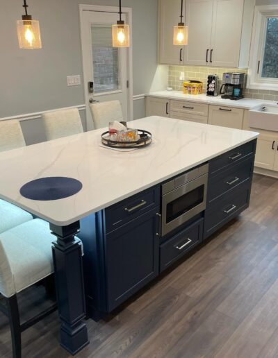 Modern kitchen with a large white island, navy blue cabinets, built-in microwave, pendant lights, and white bar stools. Door and window in the background with light gray walls.