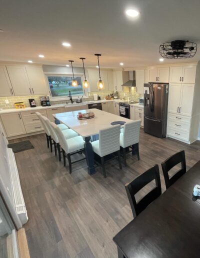 Modern kitchen with a large island and seating, pendant lights, stainless steel appliances, white cabinets, and dark wood flooring. Dining table visible in the foreground.