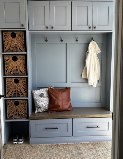 Mudroom with light blue built-in cabinets, bench, and coat hooks. Two pillows on the bench, wicker baskets in cubbies, a hanging white jacket, shoes on the floor, and a woven rug.