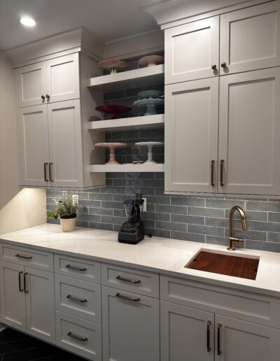 Modern kitchen with white cabinets and drawers, a built-in sink, and a countertop blender. Open shelves display colorful cake stands. Gray tiled backsplash and a potted plant enhance the decor.
