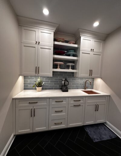 Small white kitchen nook with cabinets, drawers, and a marble countertop. Includes a sink, blender, potted plant, and shelves with dishes against a gray tiled backsplash.