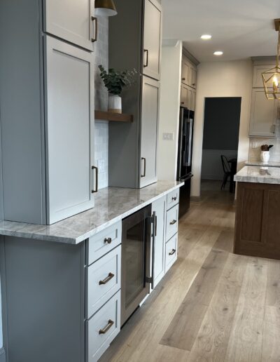 Modern kitchen with white cabinets, marble countertops, and wood flooring. A built-in oven and refrigerator are visible, along with a kitchen island and overhead lighting.