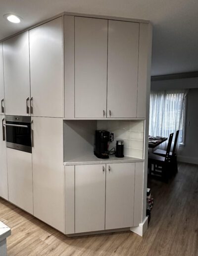 Modern kitchen with tall white cabinets, built-in oven, and a coffee maker on a countertop niche. Adjacent dining area with wooden flooring and a table near a window with sheer curtains.