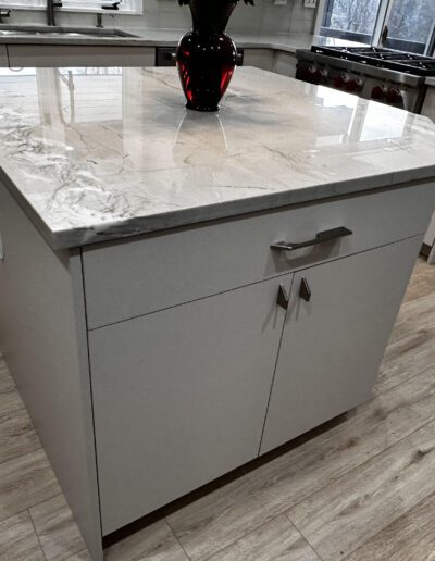 Modern kitchen island with a glossy marble countertop and white cabinets, featuring a red vase on top. The room has wooden flooring and stainless steel appliances in the background.