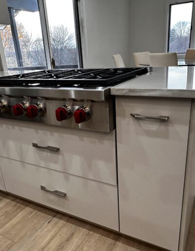 A modern kitchen with a stainless steel gas stove featuring red knobs, set in white cabinetry. Light wood flooring and a dining area with windows in the background.