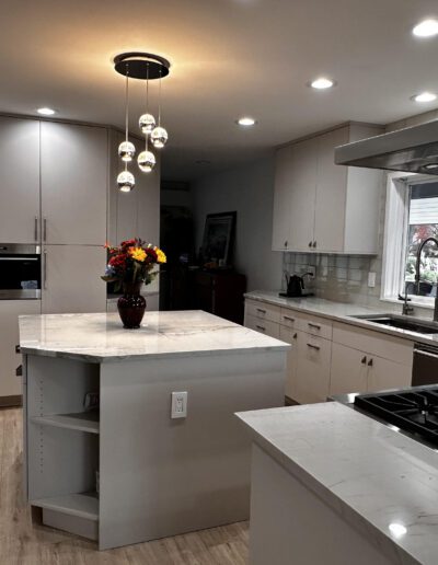 Modern kitchen with light-colored cabinets, a central island with a vase of flowers, gas stove, oven, sink, and hanging light fixture.