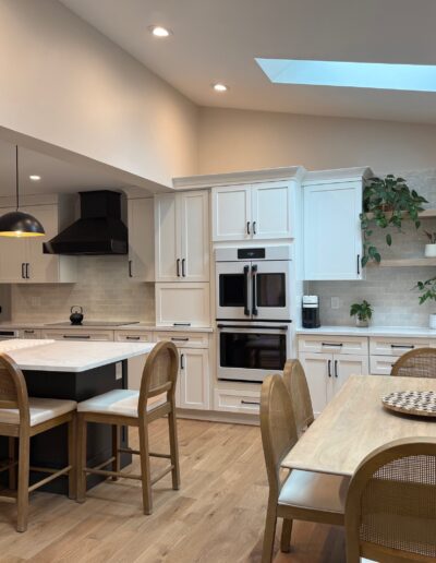 Modern kitchen with white cabinets, black accents, a central island with bar stools, a dining table with chairs, wood flooring, and a skylight above.